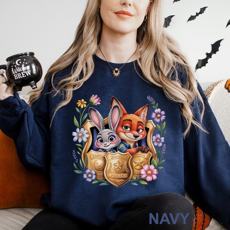 Puede incluir: Sudadera azul marino con un gr&aacute;fico colorido de Judy Hopps y Nick Wilde de Zootopia dentro de una placa de polic&iacute;a, rodeado de flores y follaje. La palabra "NAVY" est&aacute; impresa en la parte inferior.