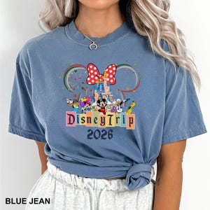 May include: Blue jean t-shirt featuring a colorful Disney Trip 2026 graphic. The design includes Mickey Mouse, Minnie Mouse, and other Disney characters, with a Ferris wheel and a large red bow. The shirt is a solid blue color.