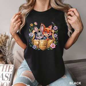 May include: Black crew neck t-shirt with a colourful graphic of Judy Hopps and Nick Wilde from Zootropolis inside a police badge, surrounded by flowers. The word "POLICE" is visible on the badge.
