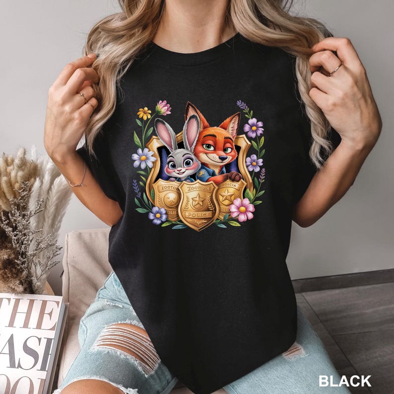 May include: Black crew neck t-shirt with a colourful graphic of Judy Hopps and Nick Wilde from Zootropolis inside a police badge, surrounded by flowers. The word "POLICE" is visible on the badge.