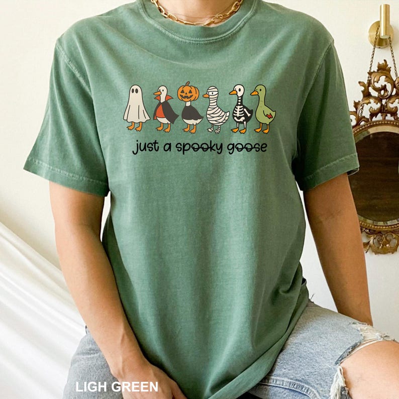 May include: Light green t-shirt with a Halloween-themed design. It features a row of cartoon geese in costumes, including a ghost, vampire, pumpkin, mummy, skeleton, and a green goose. The text "just a spooky goose" is printed below.
