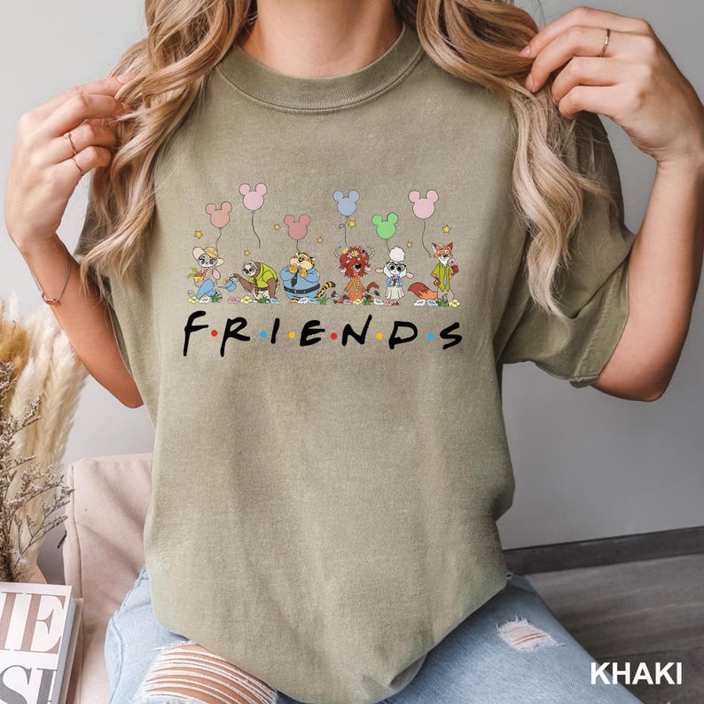 May include: Khaki t-shirt with the word "FRIENDS" in a colourful font, and cartoon animal characters holding balloon-shaped balloons above the text. The shirt is a comfortable, casual style.