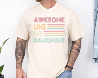 May include: Ivory-colored t-shirt with the text "AWESOME LIKE MY DAUGHTER" in a retro font. The text is layered with red, orange, yellow, teal, and green horizontal stripes. The shirt has short sleeves and a crew neck.