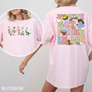 May include: A pale pink t-shirt with a vibrant graphic of characters and the text "You've Got A Friend In Me" on the back. The front of the shirt has a smaller character graphic. The t-shirt is made of a soft fabric.