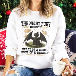 Puede incluir: Sudadera blanca con una ilustraci&oacute;n de drag&oacute;n negro y el texto "THE NIGHT FURY DRAGON" y "HEART OF A CHIEF, SOUL OF A DRAGON". El dise&ntilde;o incluye nubes, estrellas y una etiqueta de advertencia.