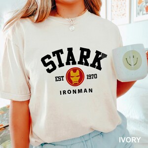 May include: White t-shirt with black text that reads "STARK EST 1970 IRONMAN" and a red and yellow Iron Man mask graphic.