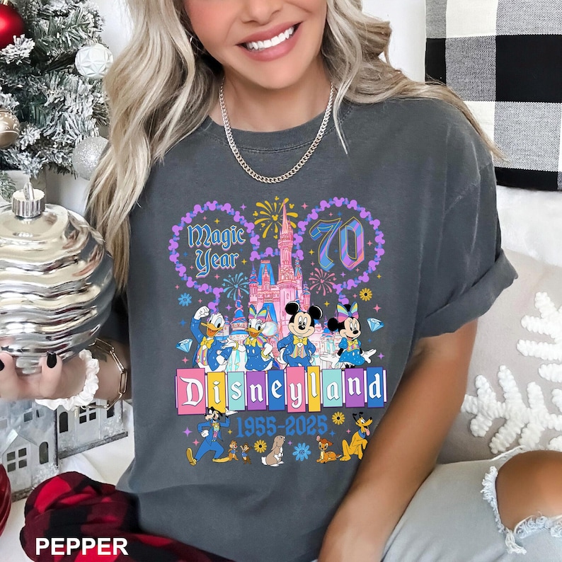 May include: Dark gray t-shirt featuring a colorful Disneyland graphic. The design includes the text "Magic Year 70" and "Disneyland 1955-2025" with cartoon characters. The shirt has short sleeves and a relaxed fit.