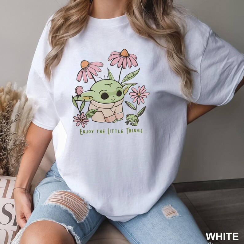 May include: White t-shirt featuring a green cartoon character, flowers, and a frog. The text "Enjoy the little things" is printed on the shirt.