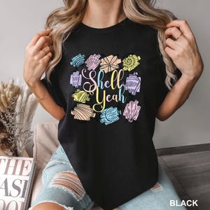 May include: Black t-shirt with a colorful seashell design and the text "Shell Yeah". The design features various pastel-colored seashells. The word "BLACK" is printed at the bottom right.