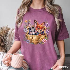 May include: Berry-coloured t-shirt featuring a graphic of Judy Hopps and Nick Wilde from Zootopia, framed by a floral design. The characters are inside police badge emblems. The word "BERRY" is printed at the bottom right.