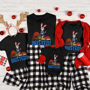 May include: A set of matching black t-shirts and a onesie with the word "BUSTED!" in blue. Each shirt features a cartoon rabbit character. The shirts are paired with black and white checkered pajama pants, creating a coordinated family outfit.