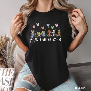 May include: Black crew neck t-shirt featuring a colorful graphic of cartoon characters and the word "FRIENDS" in a rainbow font. Above the characters are Mickey Mouse head shapes in various colors. The word "BLACK" is in the bottom right corner.