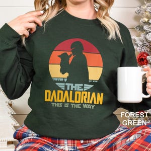 May include: A forest green long-sleeve sweatshirt featuring a graphic of a helmeted figure holding a small creature against a sunset background. The text reads THE DADALORIAN THIS IS THE WAY in yellow and light blue lettering on the front of the garment.