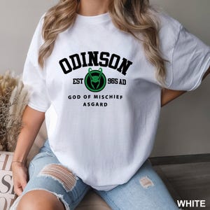 May include: White t-shirt with black text that reads "ODINSON EST 965 AD GOD OF MISCHIEF ASGARD" and a green and black logo of a horned helmet.