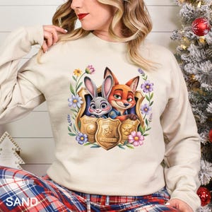 Puede incluir: Sudadera color arena con un gr&aacute;fico colorido de Judy Hopps y Nick Wilde de Zootopia, enmarcado por flores y placas de polic&iacute;a. La sudadera tiene mangas largas y cuello redondo.