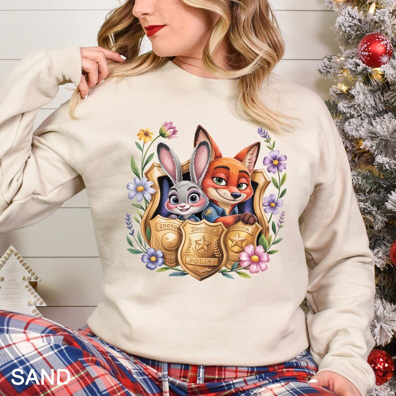 Puede incluir: Sudadera color arena con un gr&aacute;fico colorido de Judy Hopps y Nick Wilde de Zootopia, enmarcado por flores y placas de polic&iacute;a. La sudadera tiene mangas largas y cuello redondo.