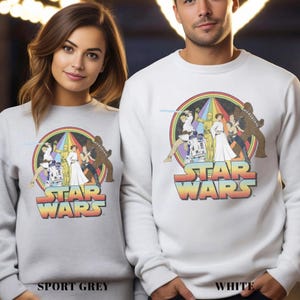 K&ouml;nnte beinhalten: Ein wei&szlig;es Sweatshirt mit einem Vintage-Star-Wars-Motiv, das Luke Skywalker, Prinzessin Leia, Han Solo, Chewbacca, R2-D2 und C-3PO zeigt. Das Motiv ist von einem Regenbogenbogen umgeben und der Text "STAR WARS" in einer Retro-Schrift.