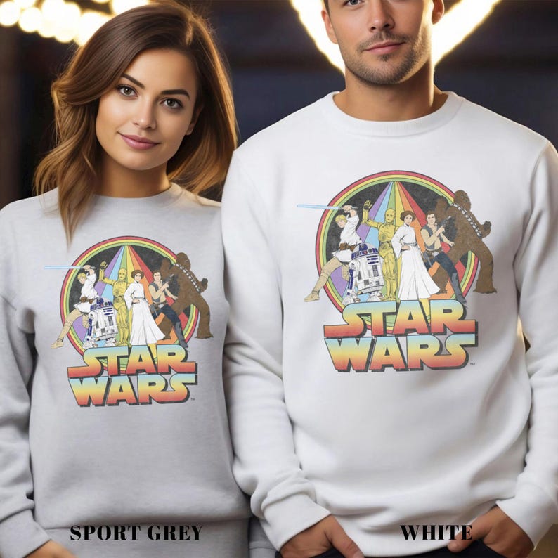 K&ouml;nnte beinhalten: Ein wei&szlig;es Sweatshirt mit einem Vintage-Star-Wars-Motiv, das Luke Skywalker, Prinzessin Leia, Han Solo, Chewbacca, R2-D2 und C-3PO zeigt. Das Motiv ist von einem Regenbogenbogen umgeben und der Text "STAR WARS" in einer Retro-Schrift.
