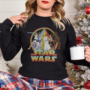 K&ouml;nnte beinhalten: Schwarzes Sweatshirt mit einem Vintage-Star-Wars-Motiv mit Figuren aus dem Originalfilm. Das Motiv zeigt Luke Skywalker, Prinzessin Leia, Han Solo, Chewbacca, R2-D2 und C-3PO. Der Text "Star Wars" ist in einem Regenbogenbogen unter den Figuren gedruckt.
