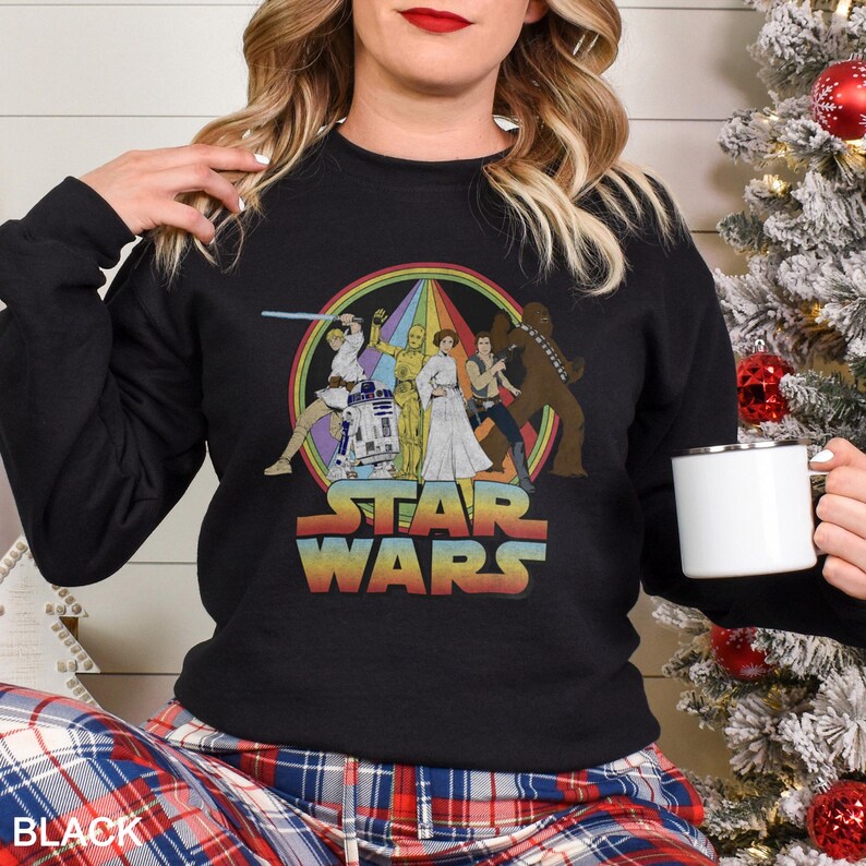 K&ouml;nnte beinhalten: Schwarzes Sweatshirt mit einem Vintage-Star-Wars-Motiv mit Figuren aus dem Originalfilm. Das Motiv zeigt Luke Skywalker, Prinzessin Leia, Han Solo, Chewbacca, R2-D2 und C-3PO. Der Text "Star Wars" ist in einem Regenbogenbogen unter den Figuren gedruckt.