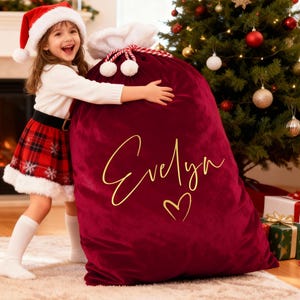 May include: A large burgundy velvet Christmas gift sack with the name "Evelyn" in gold script and a heart. The sack is tied with a white rope and white pom-poms. A child in a Santa hat and plaid skirt hugs the sack.