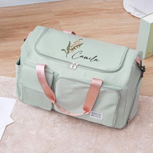 Könnte beinhalten: Hellgrüne Reisetasche mit rosa Griffen und Akzenten. Die Tasche hat zwei Vordertaschen und eine obere Klappe. Der Name "Camila" ist in Schreibschrift auf der oberen Klappe zusammen mit einem Blumendesign geschrieben. Die Tasche liegt auf einem hellen Teppich.