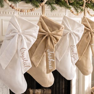 May include: Four velvet Christmas stockings hanging in a row, alternating between white and tan colours. Each stocking features a large, matching velvet bow on the front cuff, with the names Crystal, Joshua, Evelyn, and Phoebe embroidered on them.