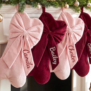 May include: Four velvet Christmas stockings in alternating light pink and deep burgundy colours. Each stocking features a large, prominent fabric bow at the top and is personalised with white embroidered names: Camila, Bailey, Chester, and Sherry.