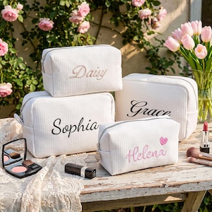 May include: Four white waffle-textured cosmetic bags with silver zippers. Each bag features embroidered names: Daisy in gold, Sophia and Grace in black, and Helena in pink with a small pink heart. The bags are arranged on a wooden surface.