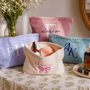 Könnte beinhalten: Vier Reißverschlusstaschen aus Kord in Lavendel, Altrosa, Creme und Blaugrün. Die lavendelfarbene Tasche hat weiße gestickte Initialen EH. Die rosafarbene Tasche zeigt ein weißes F und den Namen eleanor. Die blaugrüne Tasche hat blaue Initialen AK. Die cremefarbene Tasche zeigt den Namen clara mit einer rosa Schleife.