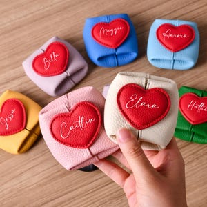 Könnte beinhalten: Eine Sammlung kleiner, quadratischer Beutel aus strukturiertem Leder in verschiedenen Farben wie Gelb, Lavendel, Blau, Hellblau, Rosa, Creme und Grün. Jeder Beutel hat einen roten herzförmigen Aufnäher mit einem in Weiß gestickten Namen.