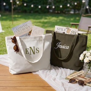 May include: A white corduroy tote bag with the embroidered green monogram ENS and a dark olive green corduroy tote bag with the embroidered white name Jessica. Both bags feature two shoulder straps and contain books and magazines.
