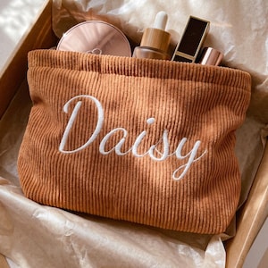 以下が含まれることがあります： 前面に白い筆記体でDaisyと刺繍された、タンカラーのコーデュロイ製ポーチ。ポーチの中には、丸いコンパクト、スポイト付きボトル、2本の長方形の口紅など、さまざまな化粧品が入っています。