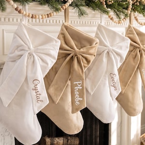 May include: Four velvet Christmas stockings in alternating white and beige colors, each featuring a large decorative bow on the front. The stockings are personalized with embroidered names including Crystal, Phoebe, Evelyn, and a fourth name.