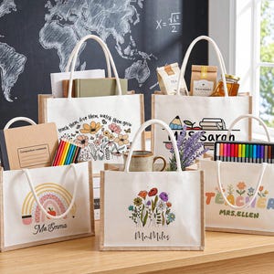 May include: Five rectangular tote bags made of white canvas with tan jute side panels and white rope handles. Each bag features unique colourful graphics including flowers, rainbows, and pencils, with personalised names like Ms. Emma and Mr. Miles.