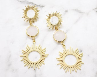 May include: A pair of gold-coloured drop earrings. Each earring has a sunburst design at the top and bottom, connected by a circular, pale pink stone. The earrings are shown against a white marble background.