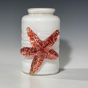 Puede incluir: Jarrón de vidrio blanco con una estrella de mar de vidrio rojo y blanco unida al lateral.