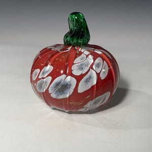 May include: A decorative glass pumpkin with a vibrant red body and white and grey accents. The pumpkin features a green stem and is a festive autumn decoration.