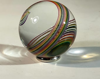Peut inclure: Une sphère en verre transparent avec des rayures colorées tourbillonnantes rouges, oranges, jaunes, vertes, bleues et violettes. La sphère repose sur une petite base argentée. Le design ressemble à un arc-en-ciel.