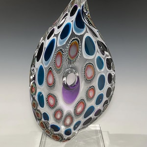 May include: A clear glass sculpture with a unique, organic shape. The sculpture features a central hole with a purple glass accent and is decorated with a pattern of white, blue, and black circles.