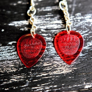 Puede incluir: Un par de pendientes de púas de guitarra rojas con cadenas plateadas. Cada púa tiene forma de corazón con un corazón más pequeño en el interior. El corazón interior tiene el texto "Jim Dunlop U.S.A." y "Stubby 1.0 mm".
