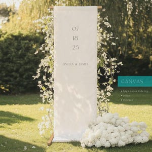 Puede incluir: Un estandarte rectangular alto de lona blanca suspendido en un marco de madera, con la fecha 07 18 25 y los nombres OVILIA & JAMES en texto gris oscuro. El estandarte est&aacute; adornado con arreglos florales blancos e incluye un cuadro de texto turquesa.