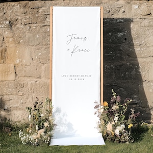 Puede incluir: Una pancarta rectangular alta de tela blanca montada sobre un marco de madera clara. La pancarta presenta el texto negro James + Krace, seguido de LULU RESORT, HAWAII y la fecha 23.12.2024. Dos grandes arreglos florales se sit&uacute;an en la base.