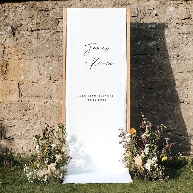 Puede incluir: Una pancarta rectangular alta de tela blanca montada sobre un marco de madera clara. La pancarta presenta el texto negro James + Krace, seguido de LULU RESORT, HAWAII y la fecha 23.12.2024. Dos grandes arreglos florales se sit&uacute;an en la base.
