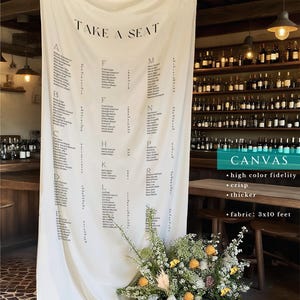 Puede incluir: Un plano de asientos de lona color crema con las palabras "TAKE A SEAT" en la parte superior. La tabla enumera los nombres alfabéticamente. El lienzo mide 91 cm por 305 cm. Un arreglo floral se encuentra en la base.