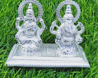 Può includere: Un set di statuette religiose argentate raffiguranti due divinità. Le figure sono sedute su una base rettangolare, con dettagli intricati e aloni circolari. Lo sfondo è verde.