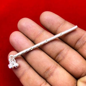 May include: A silver anklet with a linear design and small bells. The anklet features a series of small holes along its length. The bells are attached at the end of the linear design. The anklet is displayed against a red background.