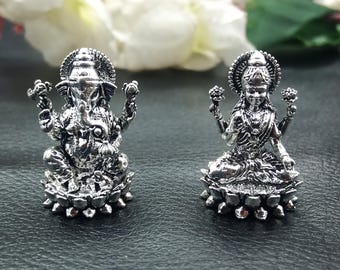 Può includere: Due statue color argento di divinità indù. Una raffigura Ganesha, il dio dalla testa di elefante, e l'altra Lakshmi, la dea della ricchezza. Entrambe sono finemente dettagliate e poggiano su basi di fiori di loto. Lo sfondo è scuro con fiori bianchi.