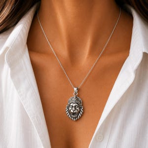 May include: A silver-toned metal necklace featuring a detailed pendant shaped like a roaring lion head. The pendant has an oxidized finish to highlight the facial features and mane. It hangs from a simple, thin silver-colored chain link necklace.