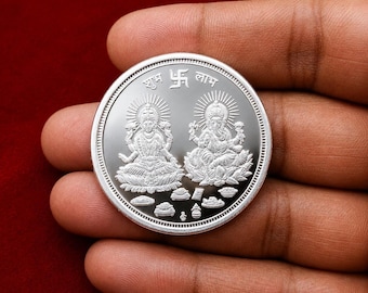 Könnte beinhalten: Eine runde Silbermünze mit geprägten Figuren von Lakshmi und Ganesha, die auf Lotusblumen sitzen. Oben in der Mitte befindet sich ein Swastika-Symbol, flankiert von dem Hindi-Text "Shubh" und "Labh". Kleine Opfergaben sind am Fuß abgebildet.